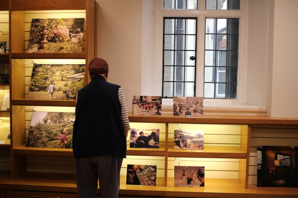 An older woman stands with her back to the camera. She looks at the photographs on display on cabinets beneath a woman. The lighting is warm.
