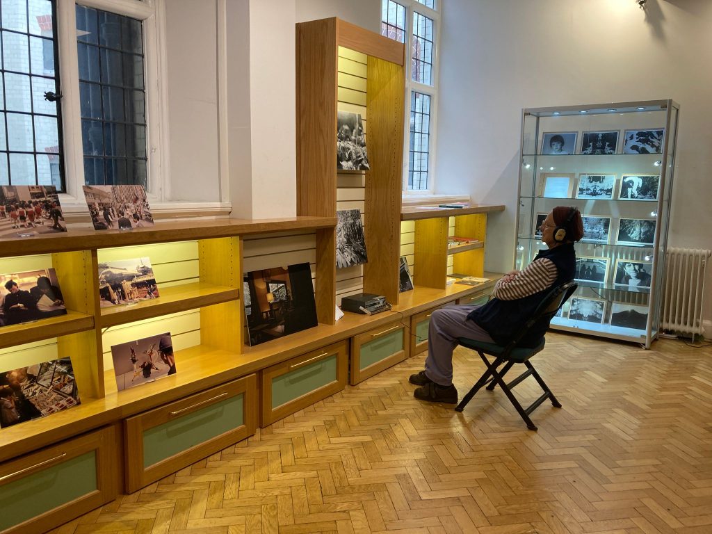 The same woman now sits on a chair in front of the images. She wears earphones and looks.